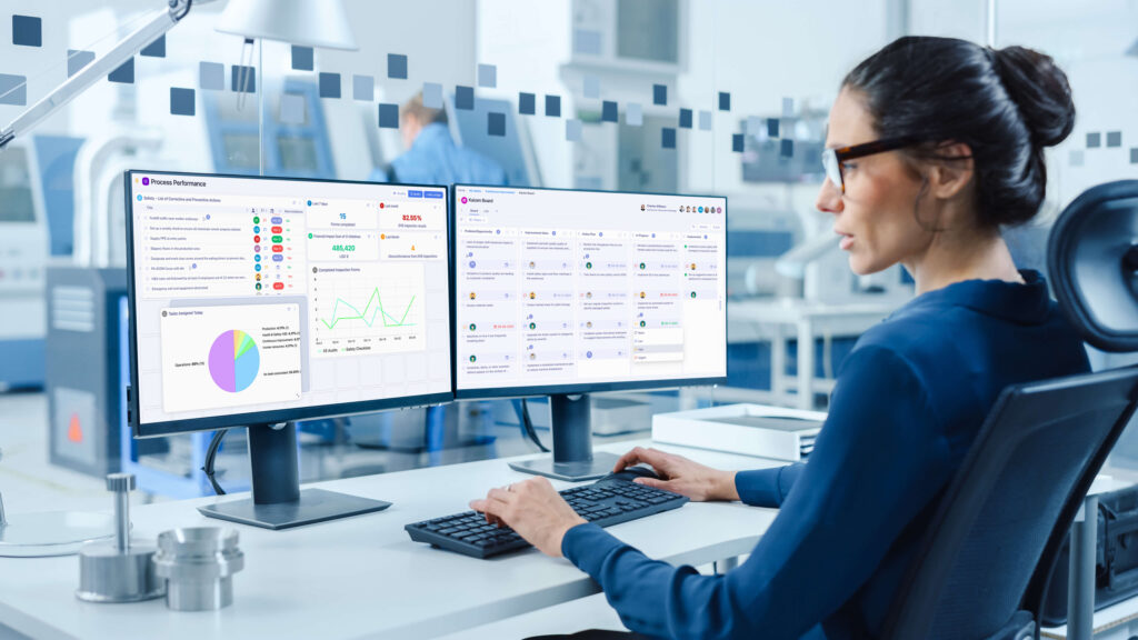 Person in an office working with dual monitors and kaizen boards, analyzing graphs and data.